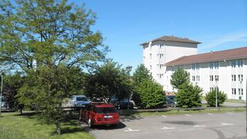 A parking lot with a red car, trees, and a white building with a red roof.