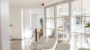 Un hall d'hôtel moderne avec des fauteuils à motifs à carreaux, une table en bois sur laquelle est posé un panier de fruits, et de grandes fenêtres décorées de guirlandes lumineuses.