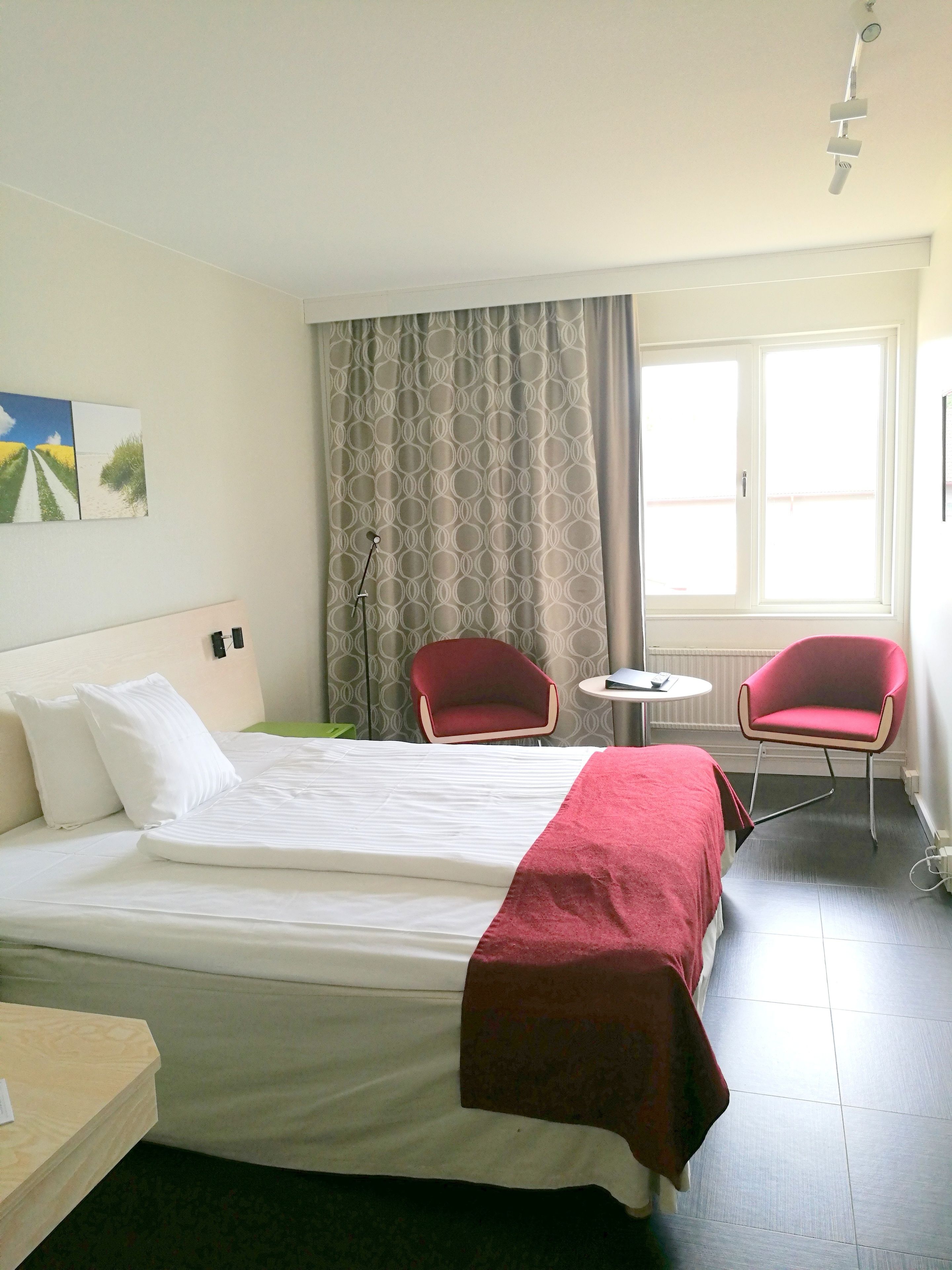 Une chambre d’hôtel avec un lit, deux chaises rouges, une petite table et une fenêtre avec des rideaux.