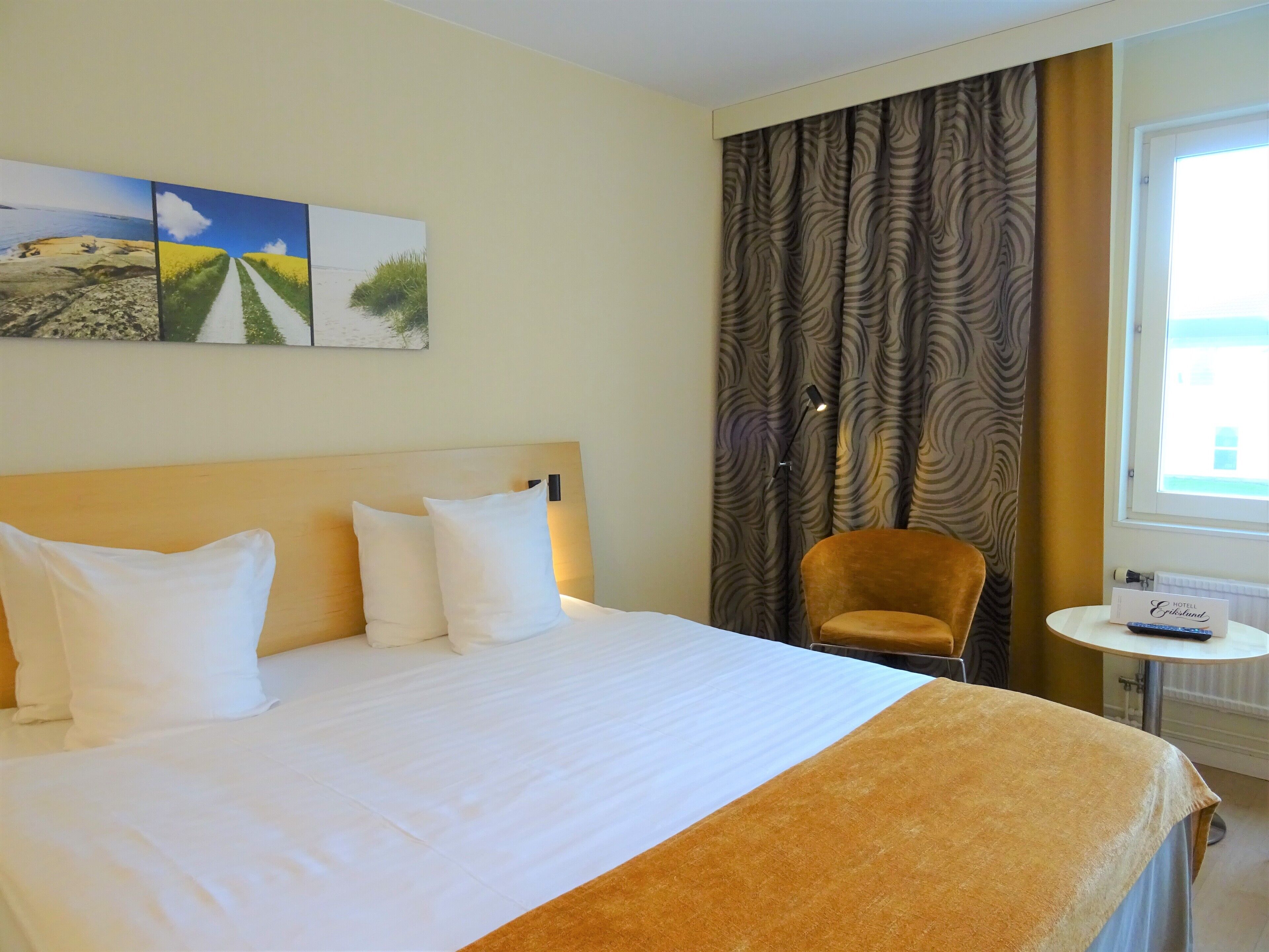Habitación de hotel con una cama grande, una silla, una mesita y un ventanal con cortinas.