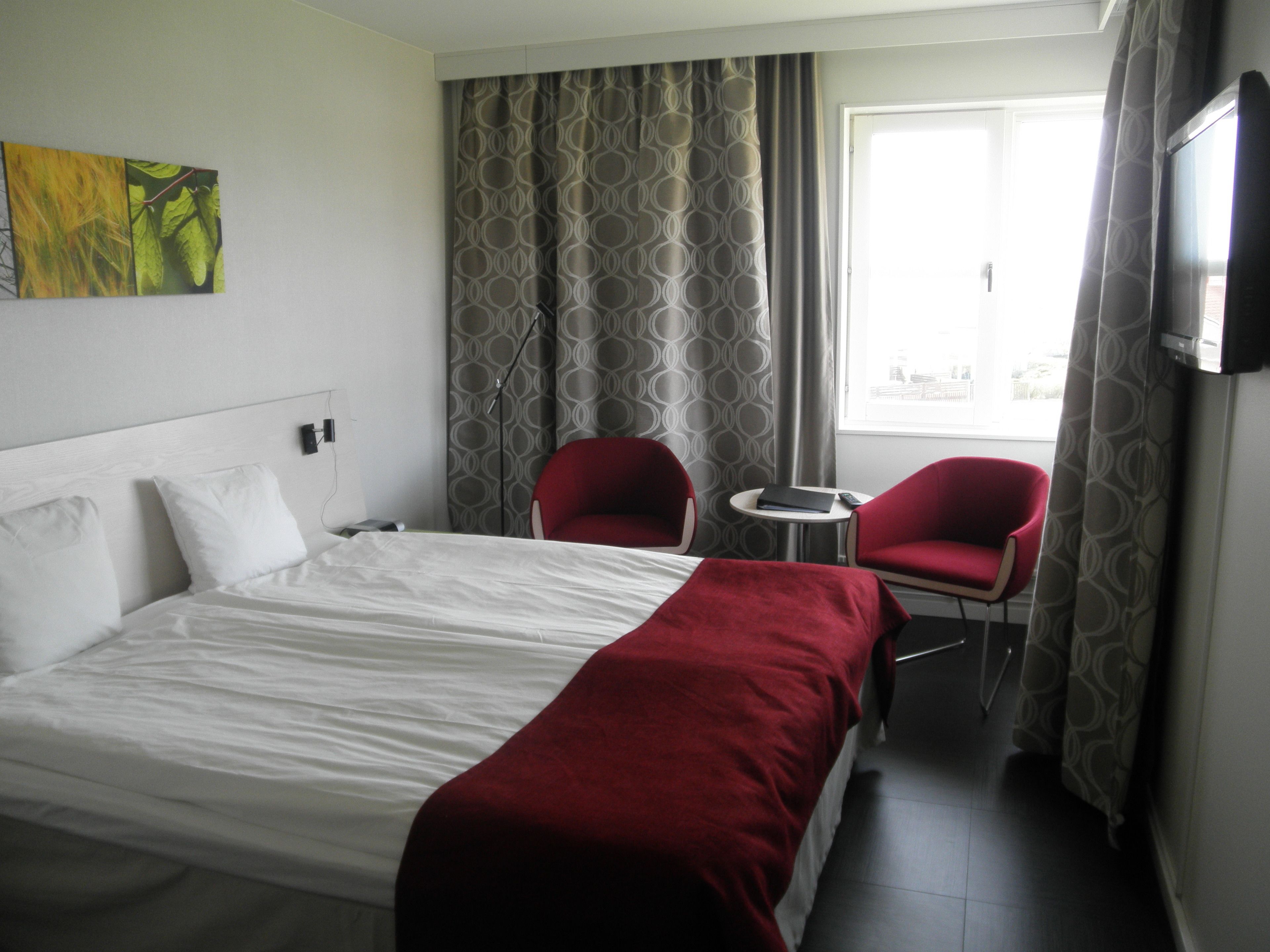 A hotel room with a bed, two red chairs, a small table, a TV, and a window with curtains.