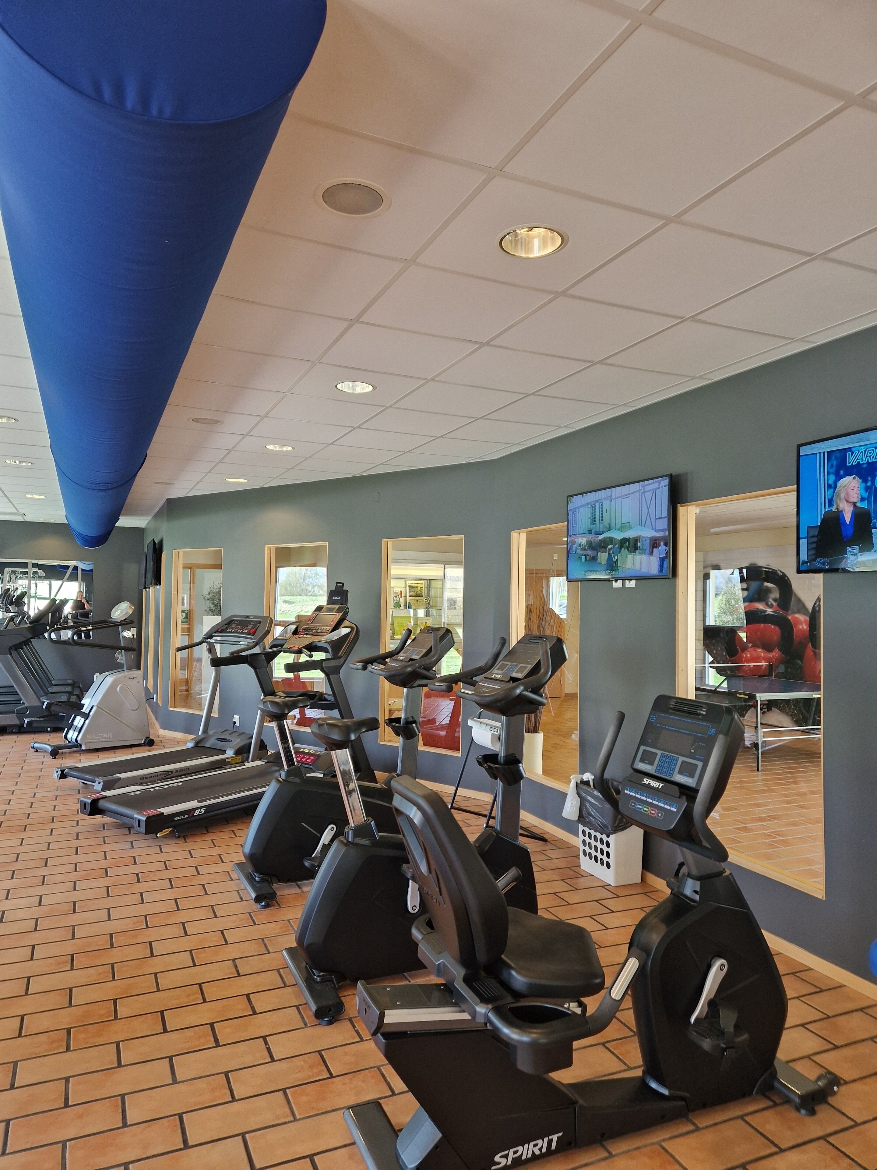 A gym with multiple treadmills and exercise bikes, a large blue ceiling feature, and a television screen displaying a news program.