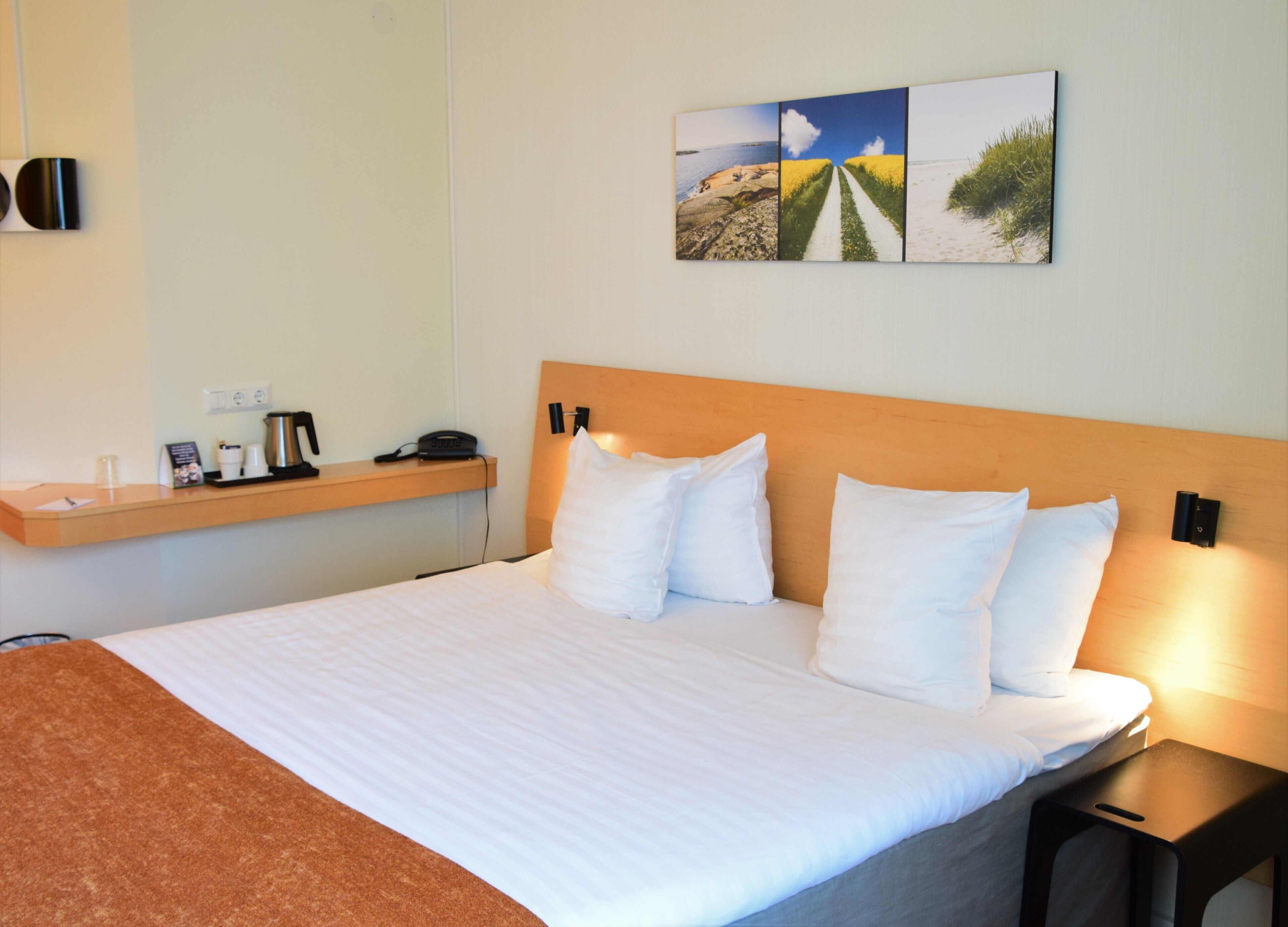 A hotel room with a large bed, a wooden headboard, a bedside table with a lamp, a shelf with a phone, a coffee maker, and a framed picture on the wall.