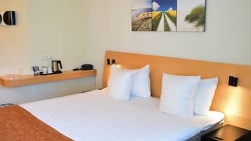 Habitación de hotel con una cama grande, cabecera de madera, mesita de noche con lámpara, estante con teléfono, cafetera y un cuadro en la pared.