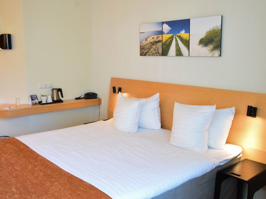 Habitación de hotel con una cama grande, cabecera de madera, mesita de noche con lámpara, estante con teléfono, cafetera y un cuadro en la pared.
