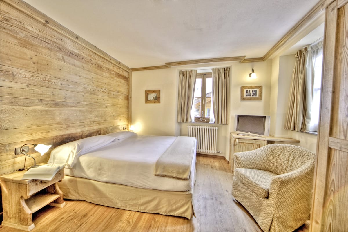 A bedroom with a wooden headboard, a bed with white linens, a bedside table with a lamp, a chair, and a TV on a stand.
