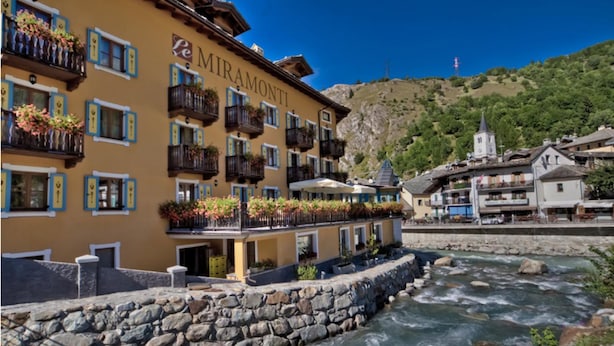 A hotel with balconies adorned with flowers, a stone wall in front, and a river nearby. - Le Miramonti Hotel & Wellness (La Thuile)