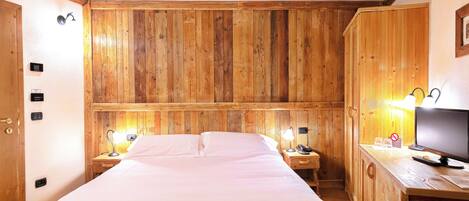 A wooden-paneled room with a bed, bedside lamps, a desk with a computer, and a television.