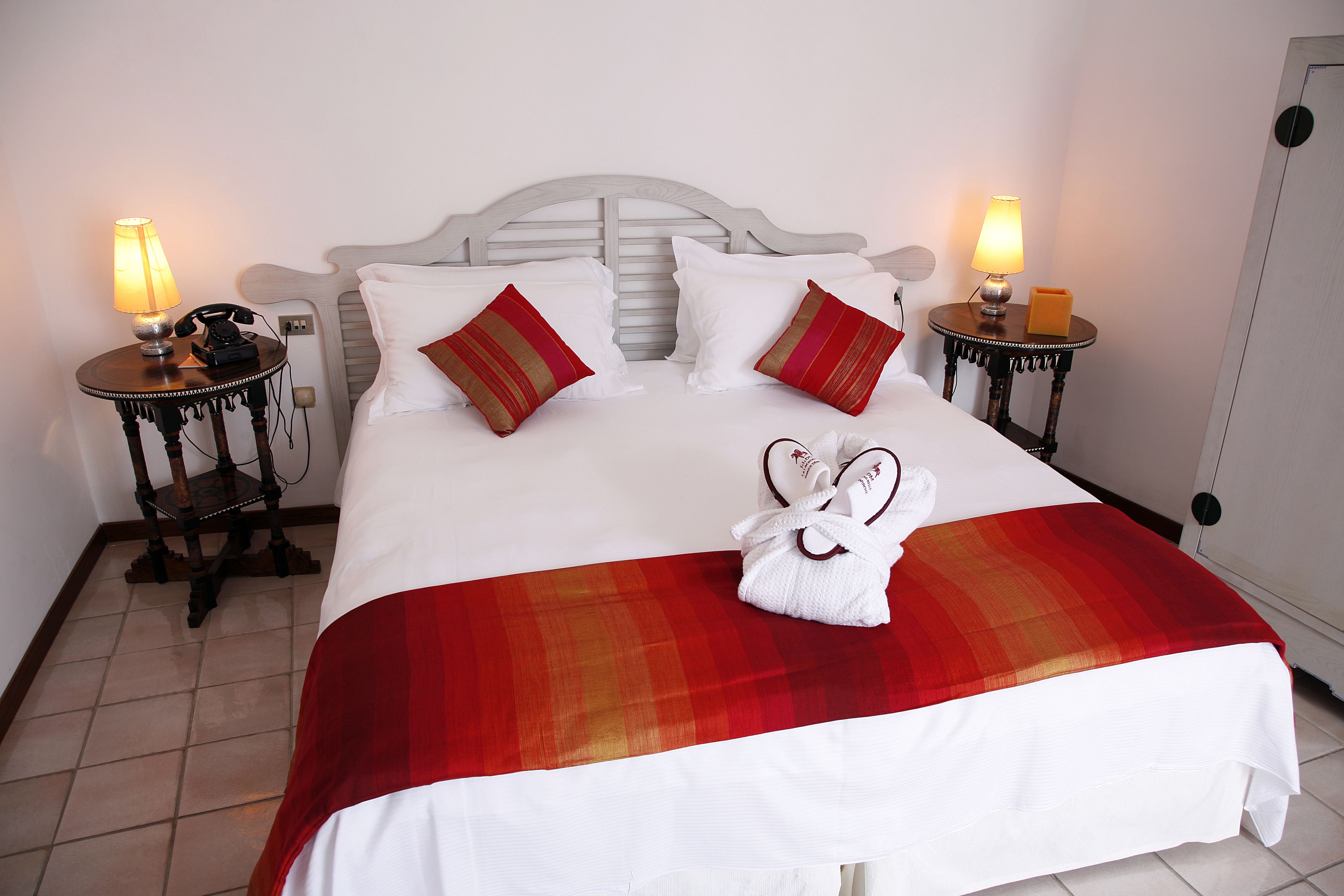 A neatly made bed with a red and white blanket, two bedside lamps, a telephone, and a pair of slippers on the floor.