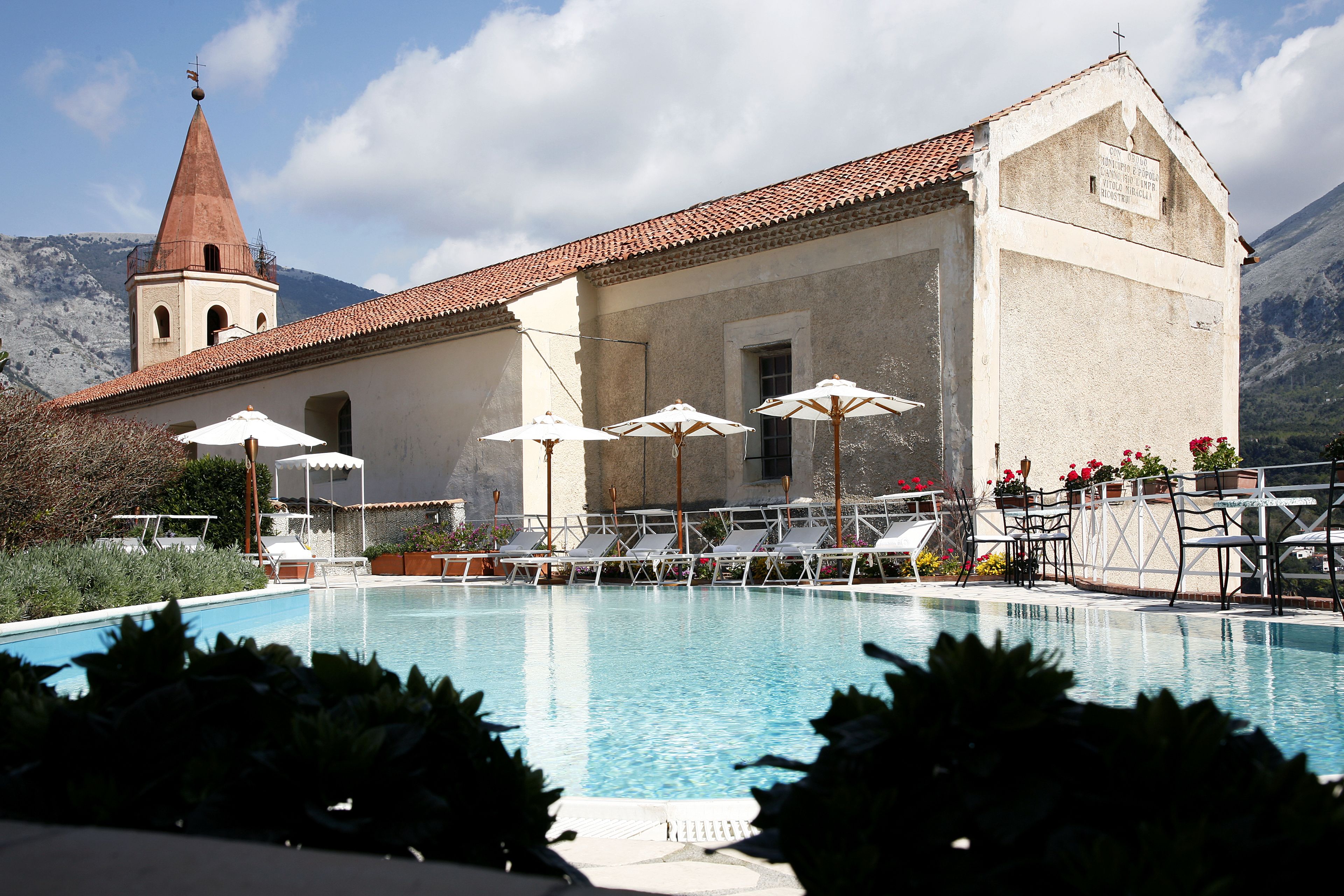A swimming pool with lounge chairs and umbrellas, a church with a bell tower, and mountains in the background.