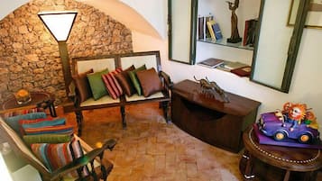 A cozy living room with a stone wall, a wooden bench with cushions, a small table with a lamp, and a bookshelf with books and a statue.