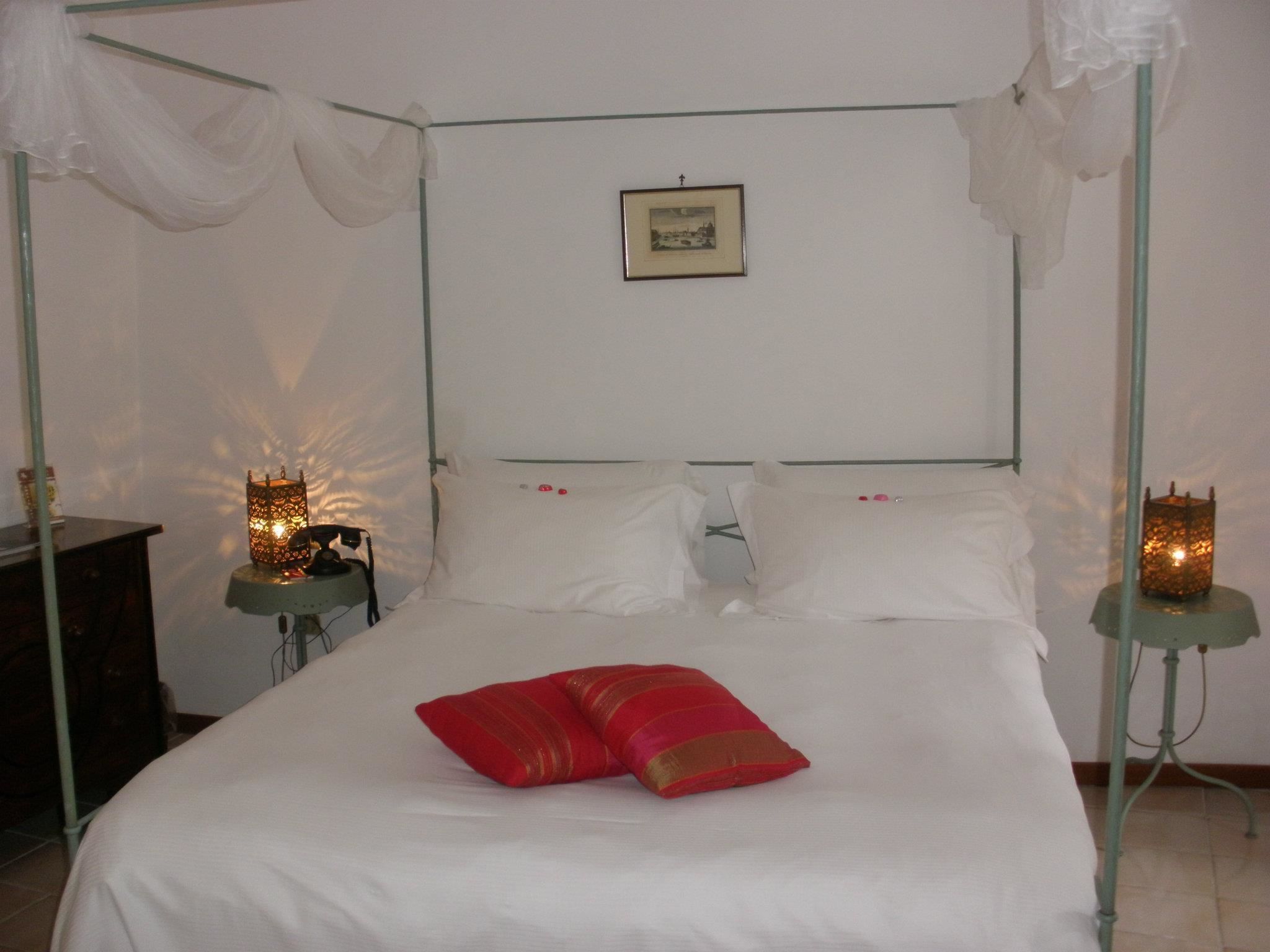 A four-poster bed with white linens and red pillows, two bedside tables with candles, and a framed picture on the wall.