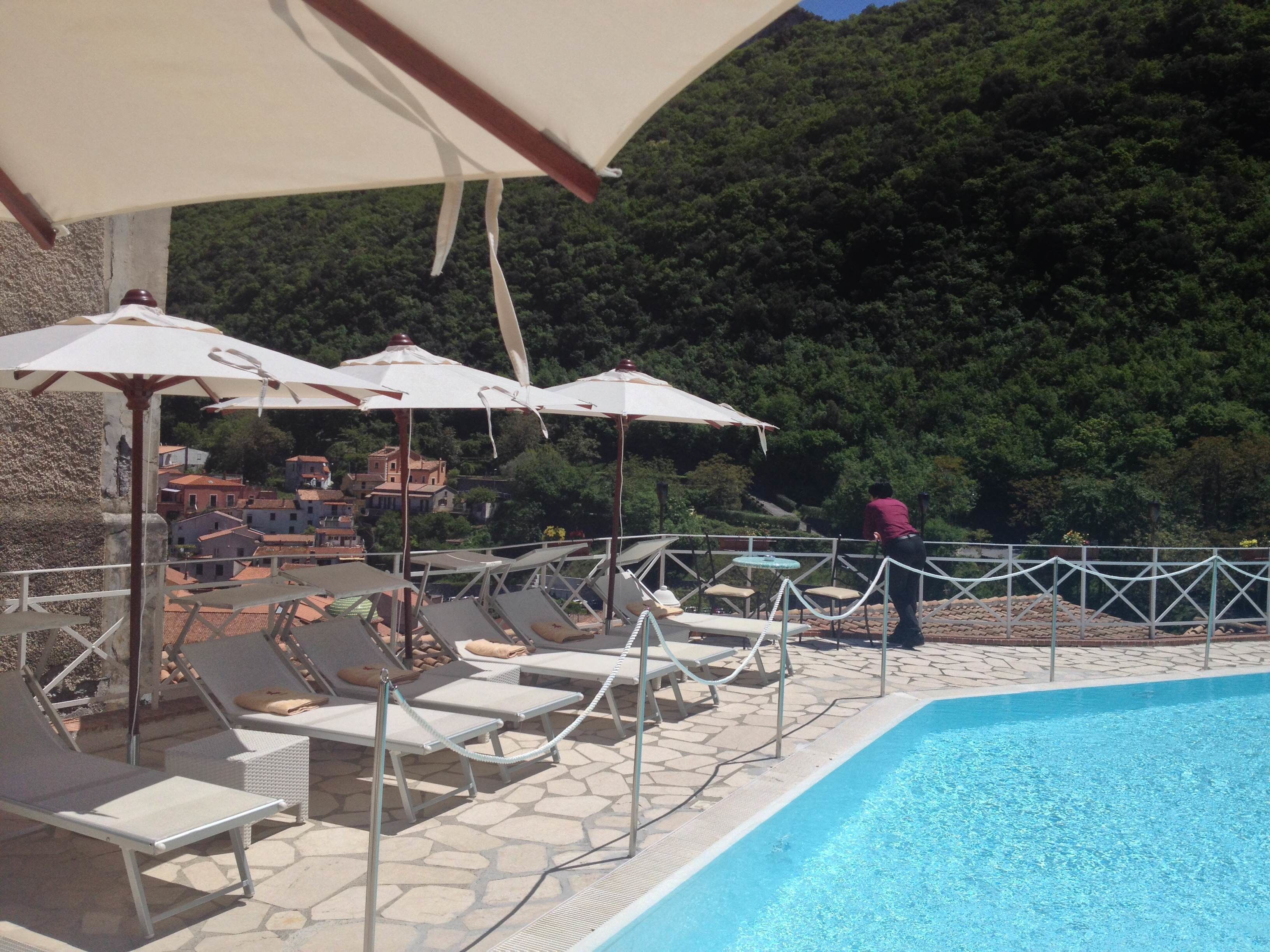 A poolside area with lounge chairs and umbrellas, surrounded by a stone deck and a view of a hillside with buildings.