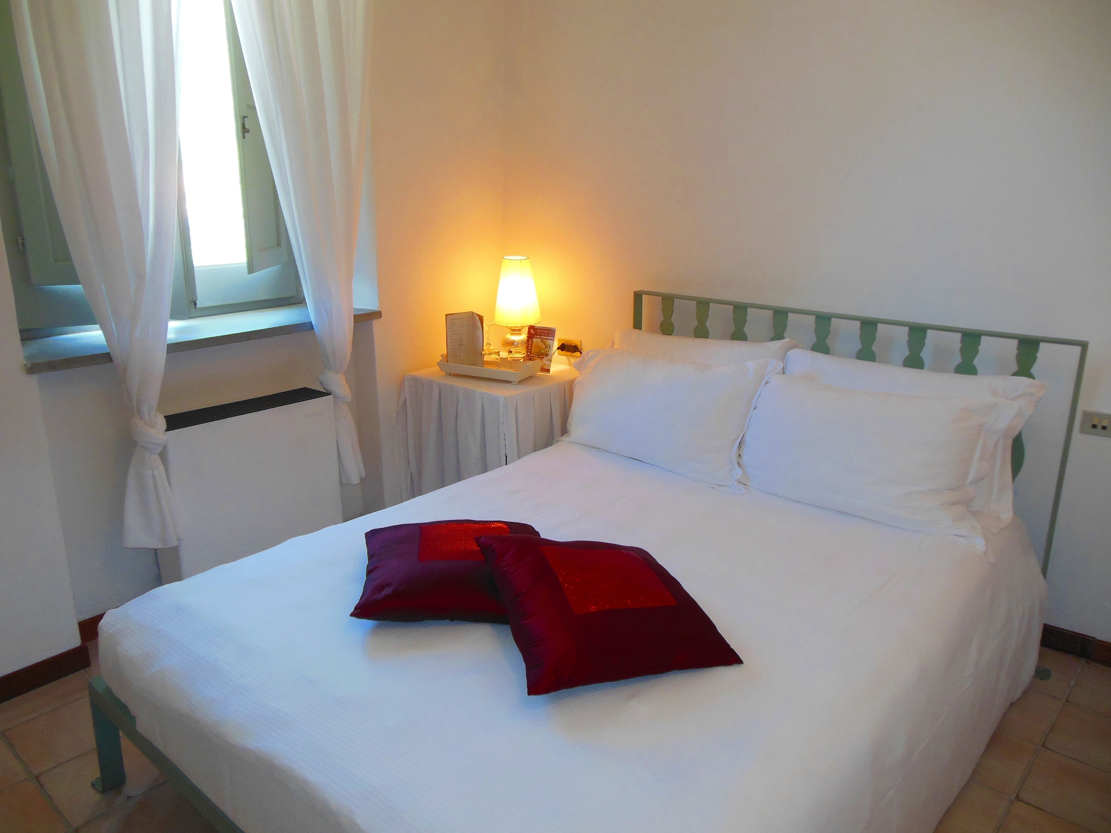 A neatly made bed with white linens and two red pillows, a bedside table with a lamp, and a window with white curtains.