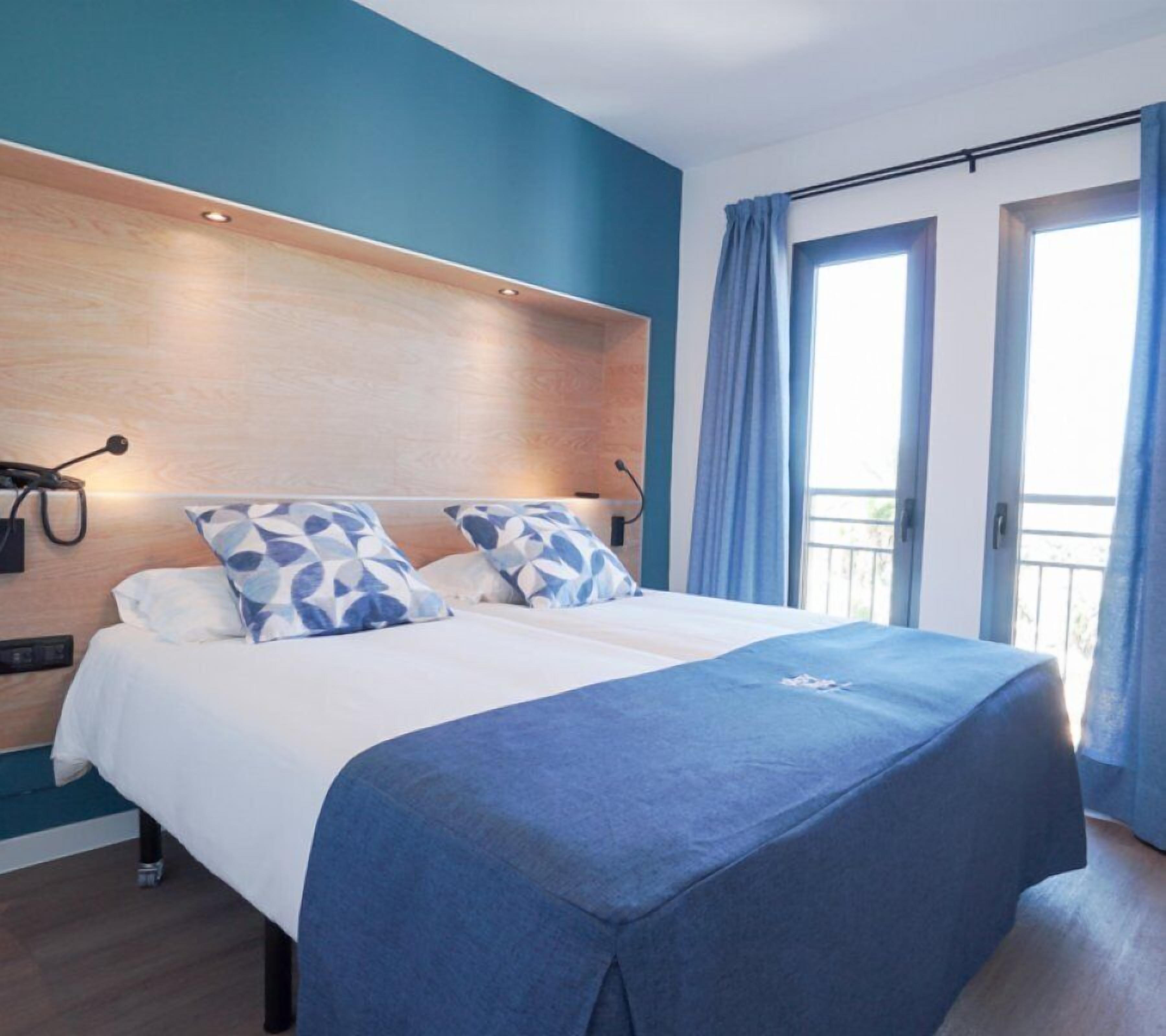 A hotel room with a bed, a wooden headboard, a nightstand, a desk, and a window with curtains.