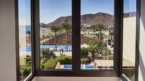 Vista desde una ventana de un resort, con piscina, palmeras y una playa al fondo.