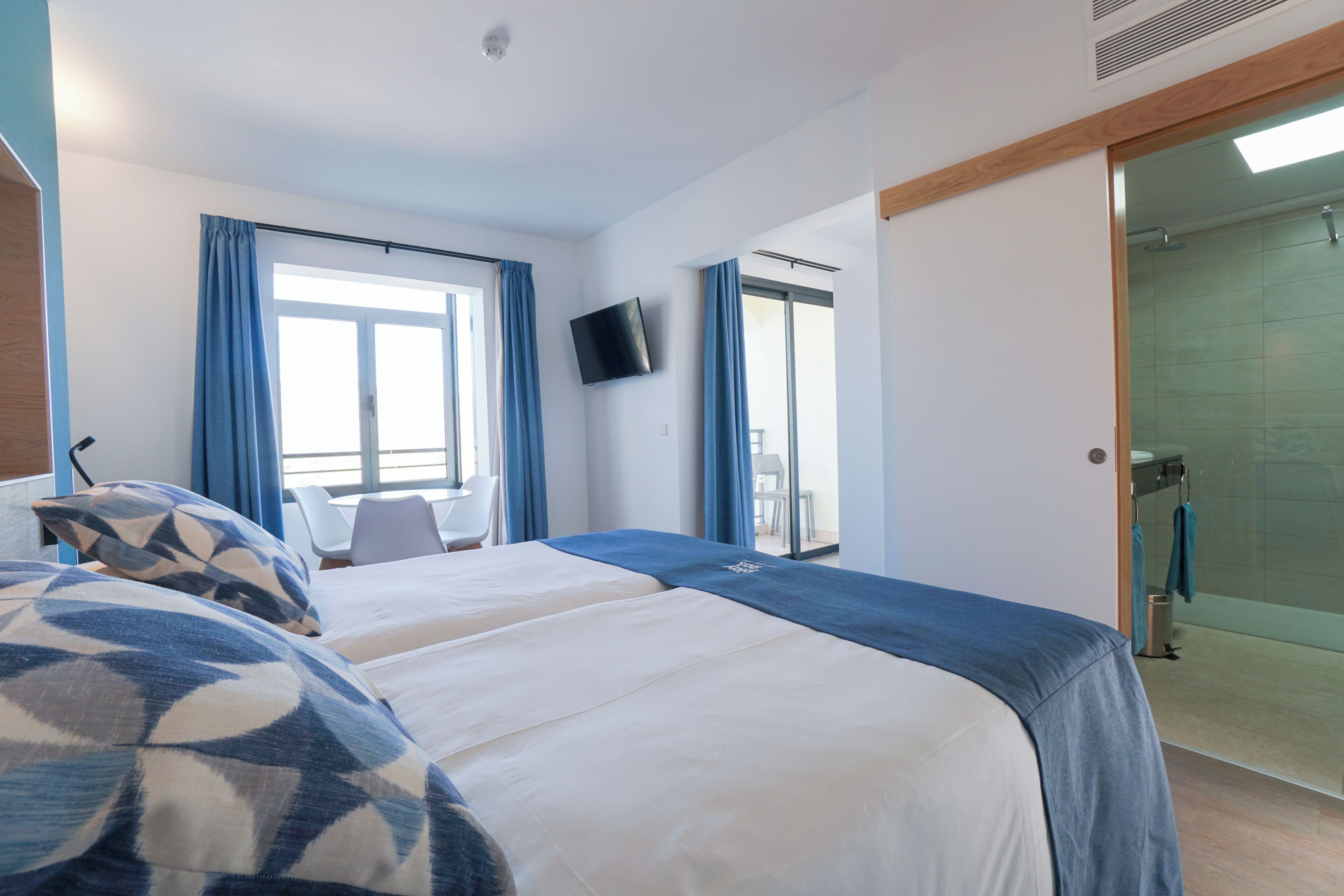 A hotel room with two beds, a TV, a shower area, and a balcony with blue curtains.