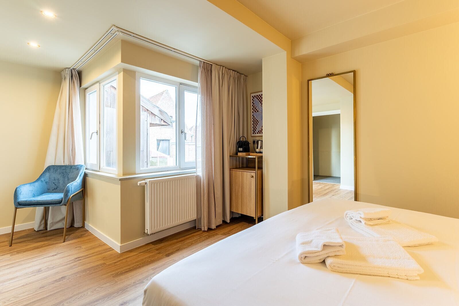 A hotel room with a bed, a chair, a radiator, a window with curtains, a mirror, and a small table with a handbag.