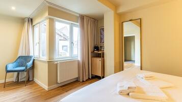 Habitación de hotel con cama, una silla, un radiador, ventana con cortinas, un espejo y una mesita con un bolso.