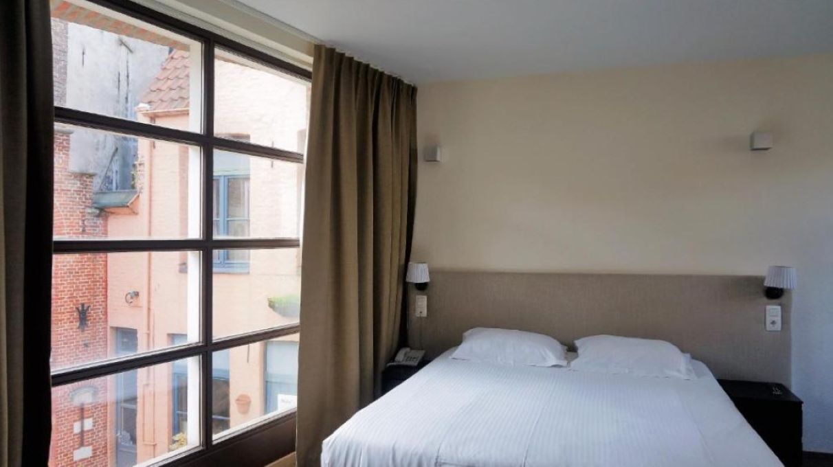 Une chambre d’hôtel avec une grande fenêtre, un lit, deux tables de chevet et une lampe fixée au mur.