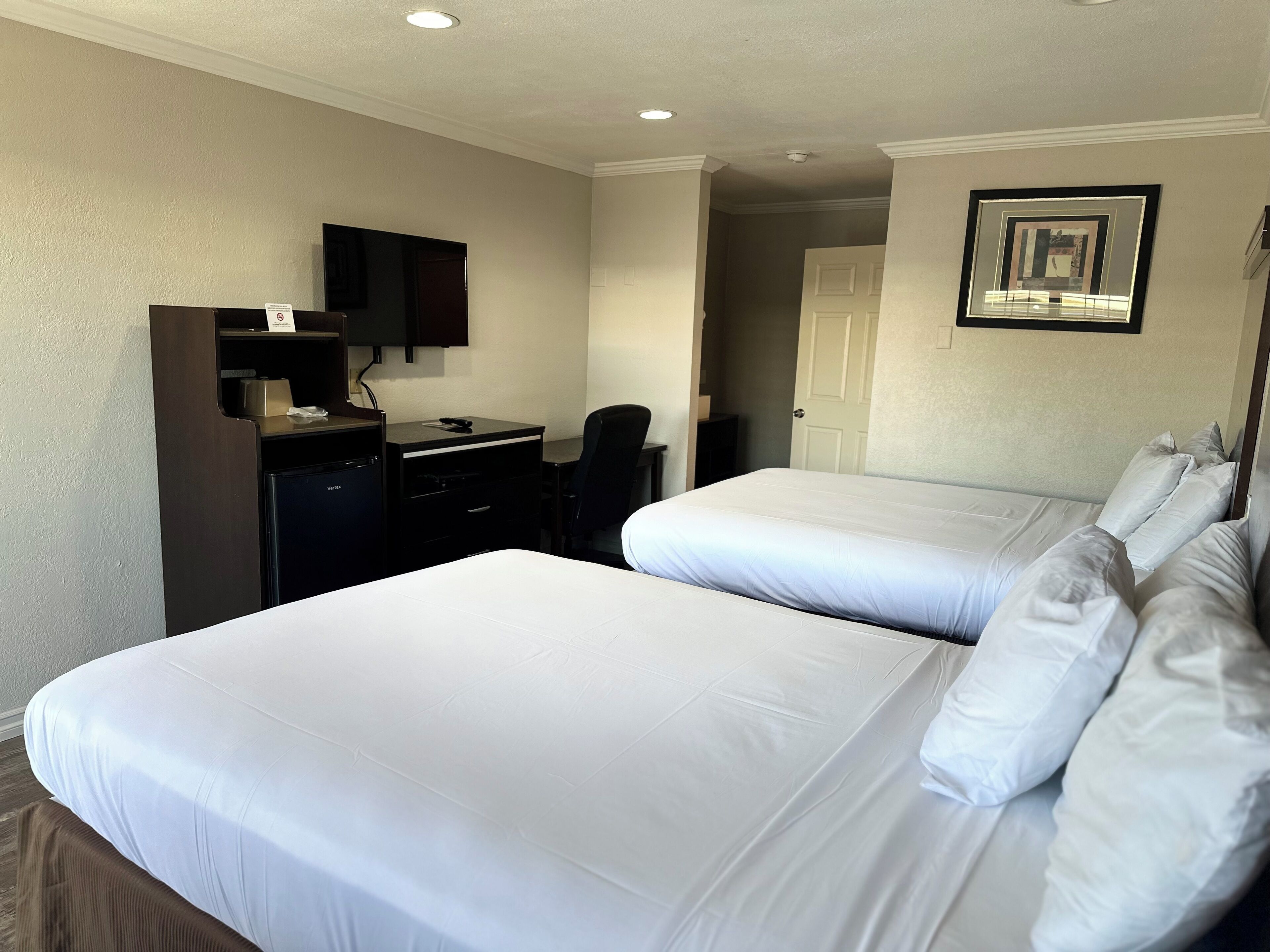 Una habitación de hotel con dos camas, un escritorio con una silla, un televisor y una puerta que da a otra habitación.