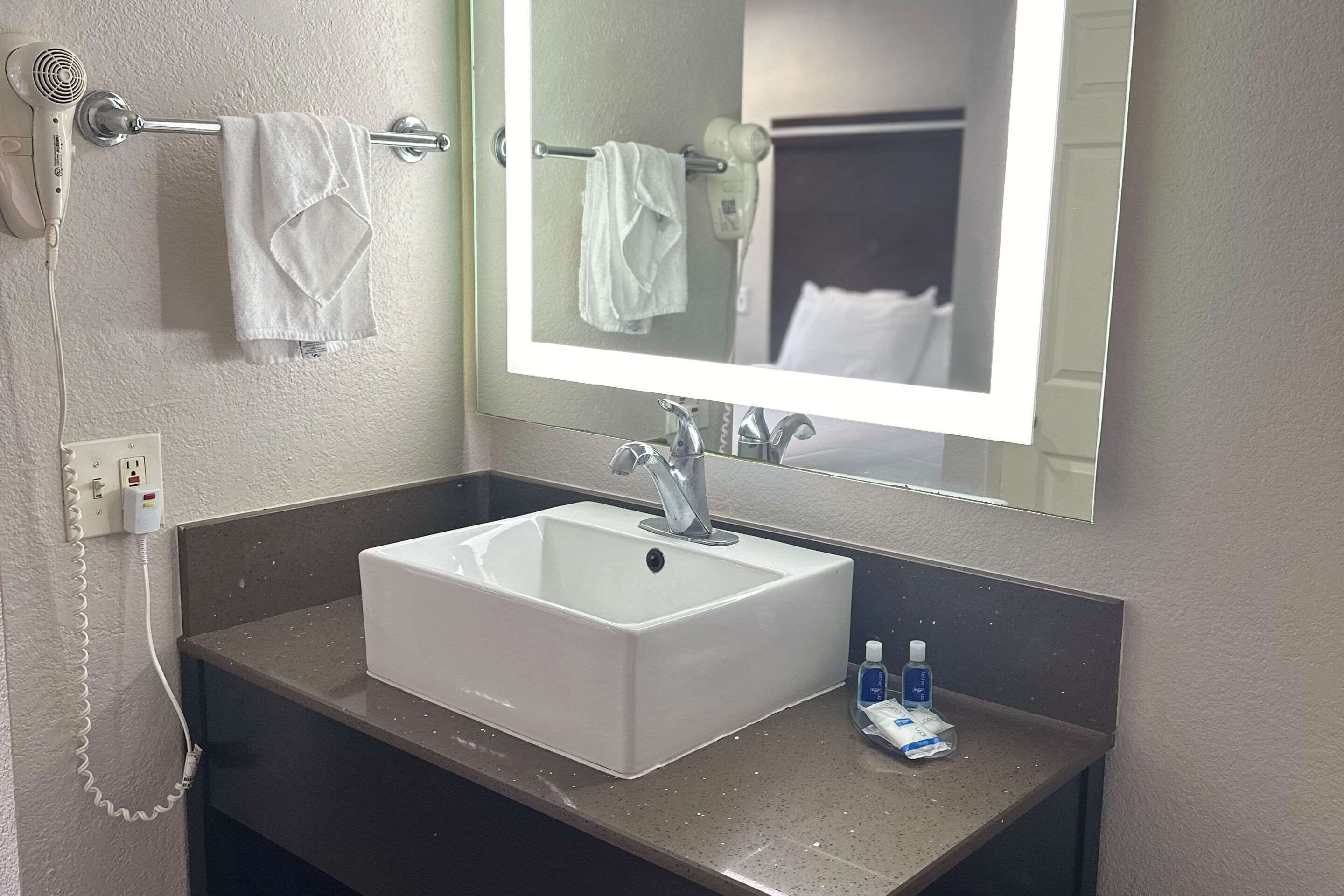 Une salle de bain avec un lavabo blanc, un miroir, un porte-serviettes avec deux serviettes, un sèche-cheveux et un flacon de gel hydroalcoolique sur le comptoir.