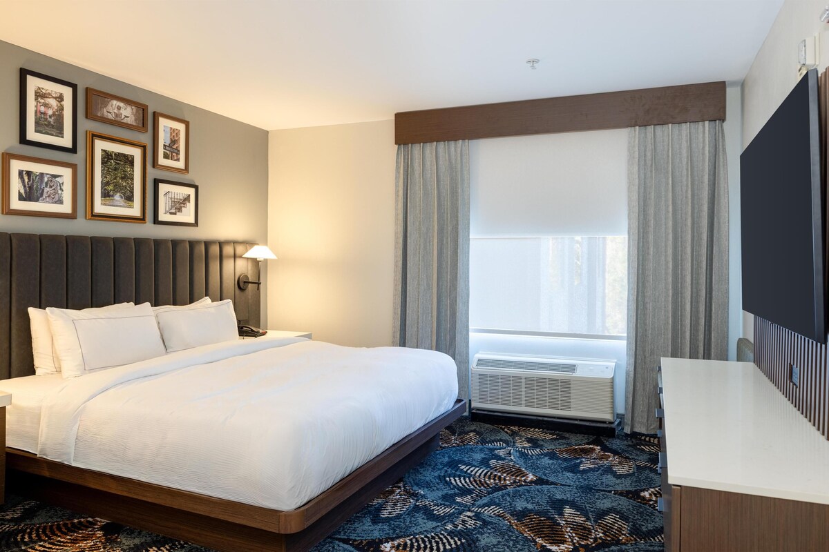 A hotel room with a large bed, a desk, a flat-screen TV, a window with curtains, and framed pictures on the wall.