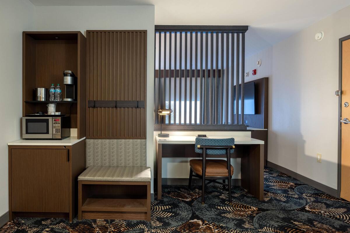 A compact hotel room with a kitchenette, a desk, a chair, a patterned carpet, and a window with blinds.