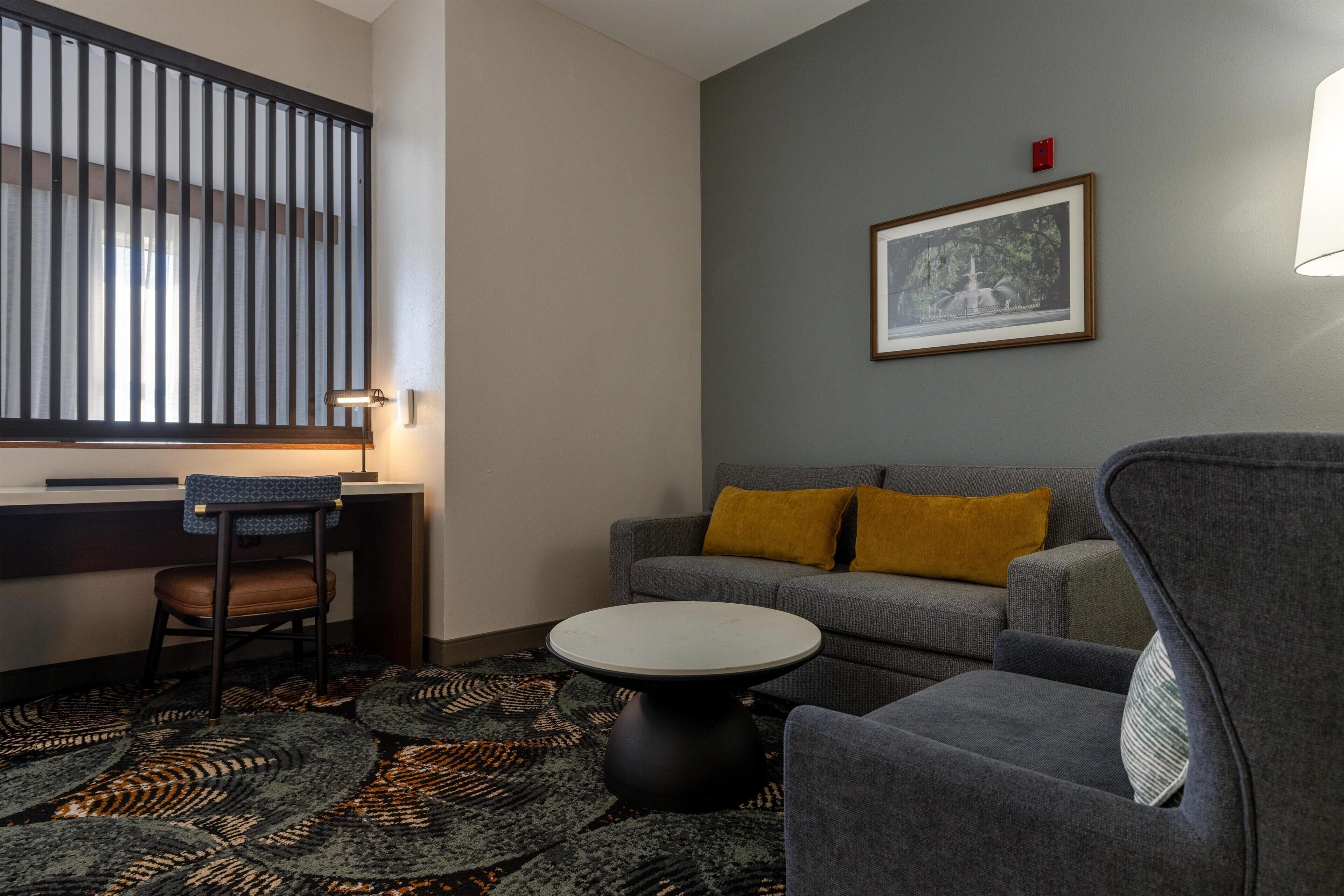 Une pièce avec un canapé gris, une table basse ronde, un tapis à motifs, un tableau encadré au mur et une fenêtre avec des stores.