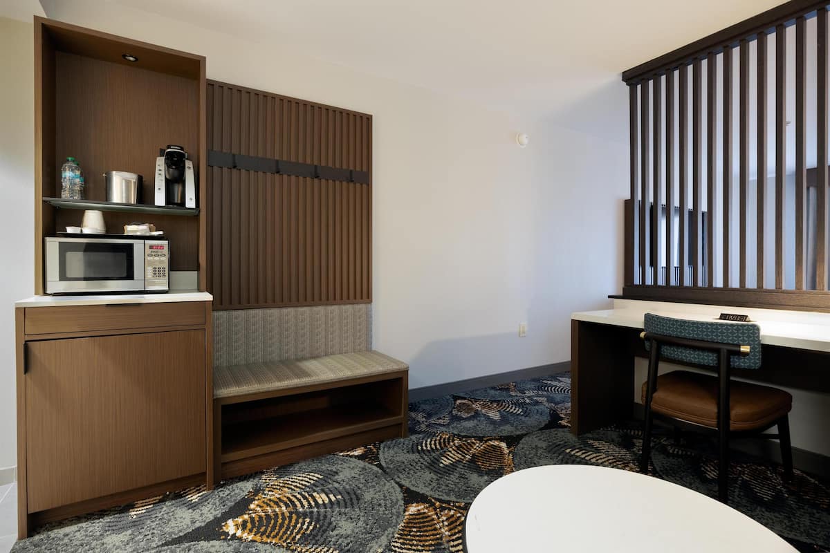 A compact hotel room with a kitchenette, a microwave, a coffee maker, and a seating area with a sofa and a chair.