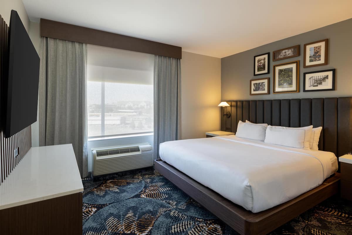 A hotel room with a bed, a desk, a TV, a window with curtains, a wall with framed pictures, and a patterned carpet.
