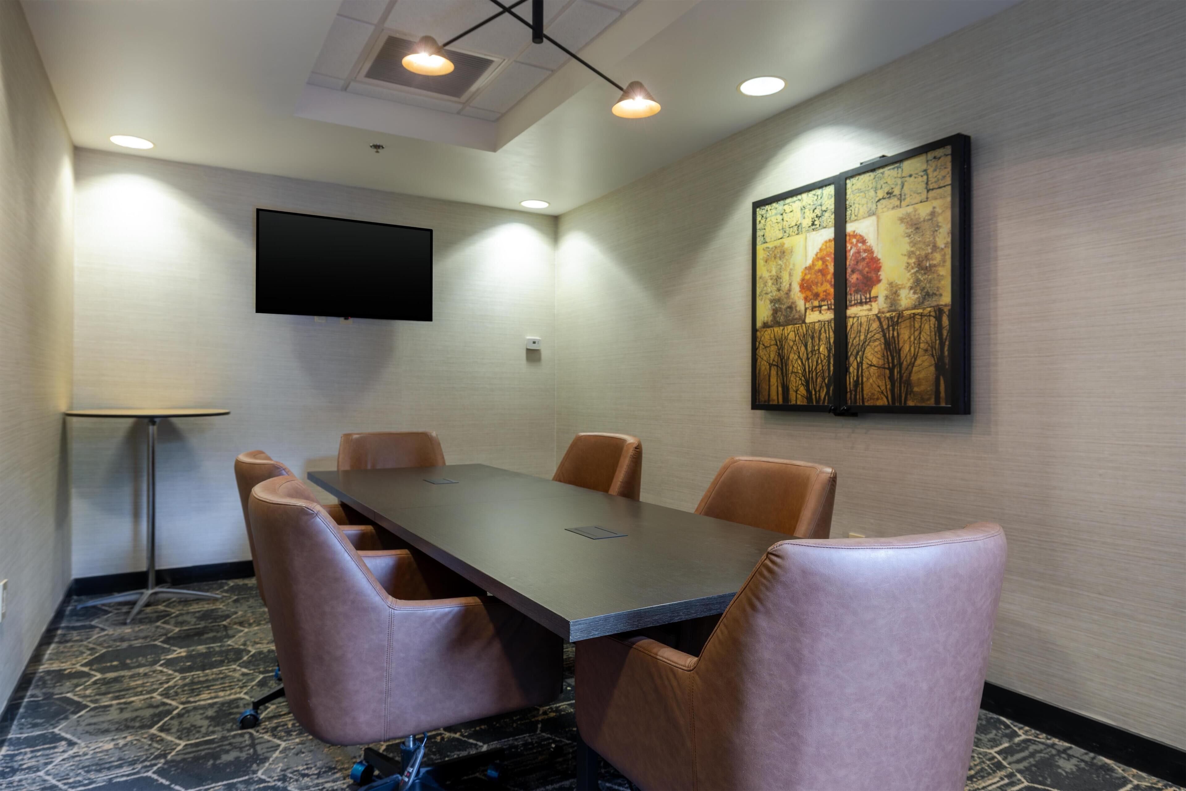Une salle de conférence avec une longue table, six chaises, un téléviseur à écran plat fixé au mur et un tableau au-dessus de la table.