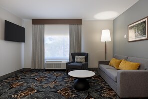 Une chambre d’hôtel équipée d’un téléviseur à écran plat, d’un canapé, d’un fauteuil, d’une table basse ronde, d’une lampe et d’une fenêtre avec des rideaux.
