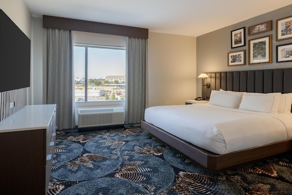 A hotel room with a large bed, a desk, a window with curtains, and framed pictures on the wall.