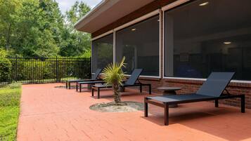 A patio with lounge chairs and a small palm tree, a brick wall, and a glass door.