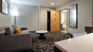 Een hotelkamer met een grijze bank, een rond tafeltje, een kamerbreed tapijt met dessin, en een spiegel aan de muur.