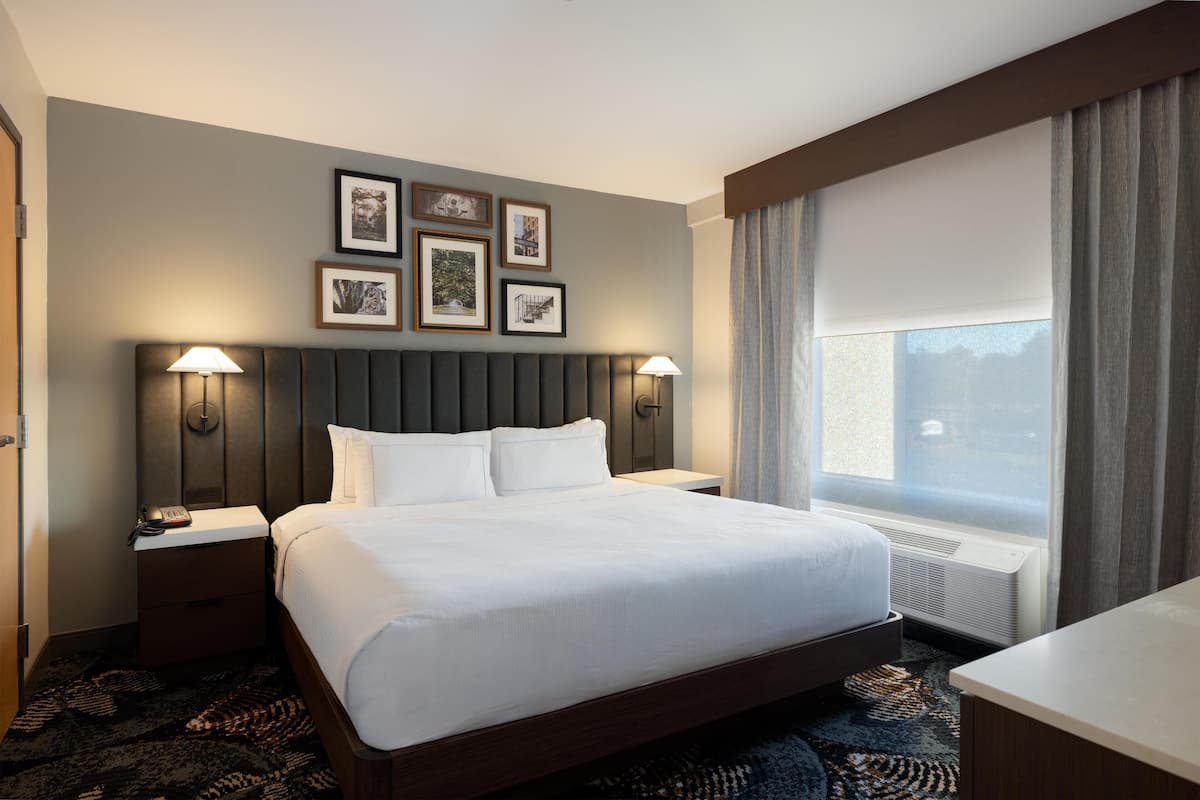 A hotel room with a large bed, two bedside tables with lamps, a headboard, framed pictures on the wall, a window with curtains, and a carpeted floor.