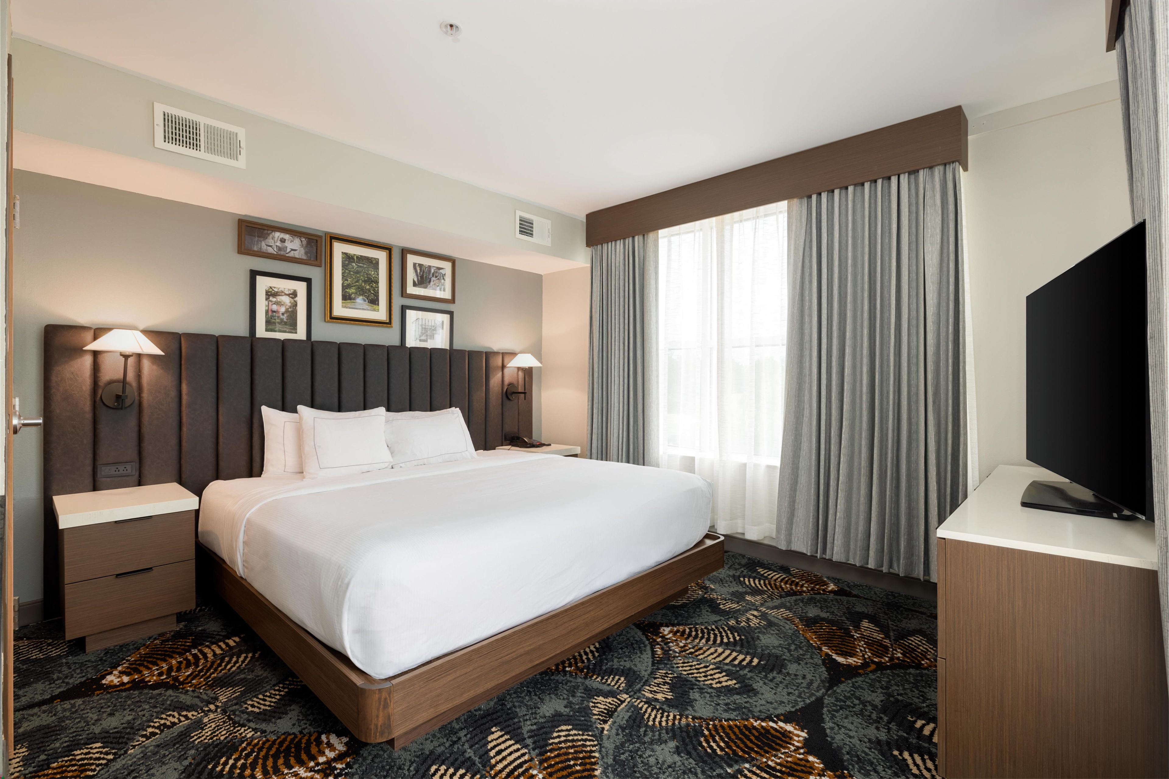 Une chambre d’hôtel avec un grand lit, deux tables de chevet, un téléviseur à écran plat, une fenêtre avec des rideaux et des tableaux encadrés accrochés au mur.