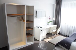 Una camera d'albergo moderna con cabina armadio, una scrivania con sedia, un tavolino con lampada e un letto.
