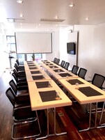 Una sala conferenze con lunghe tavolate, sedie, uno schermo per proiettore e un televisore appeso al muro.