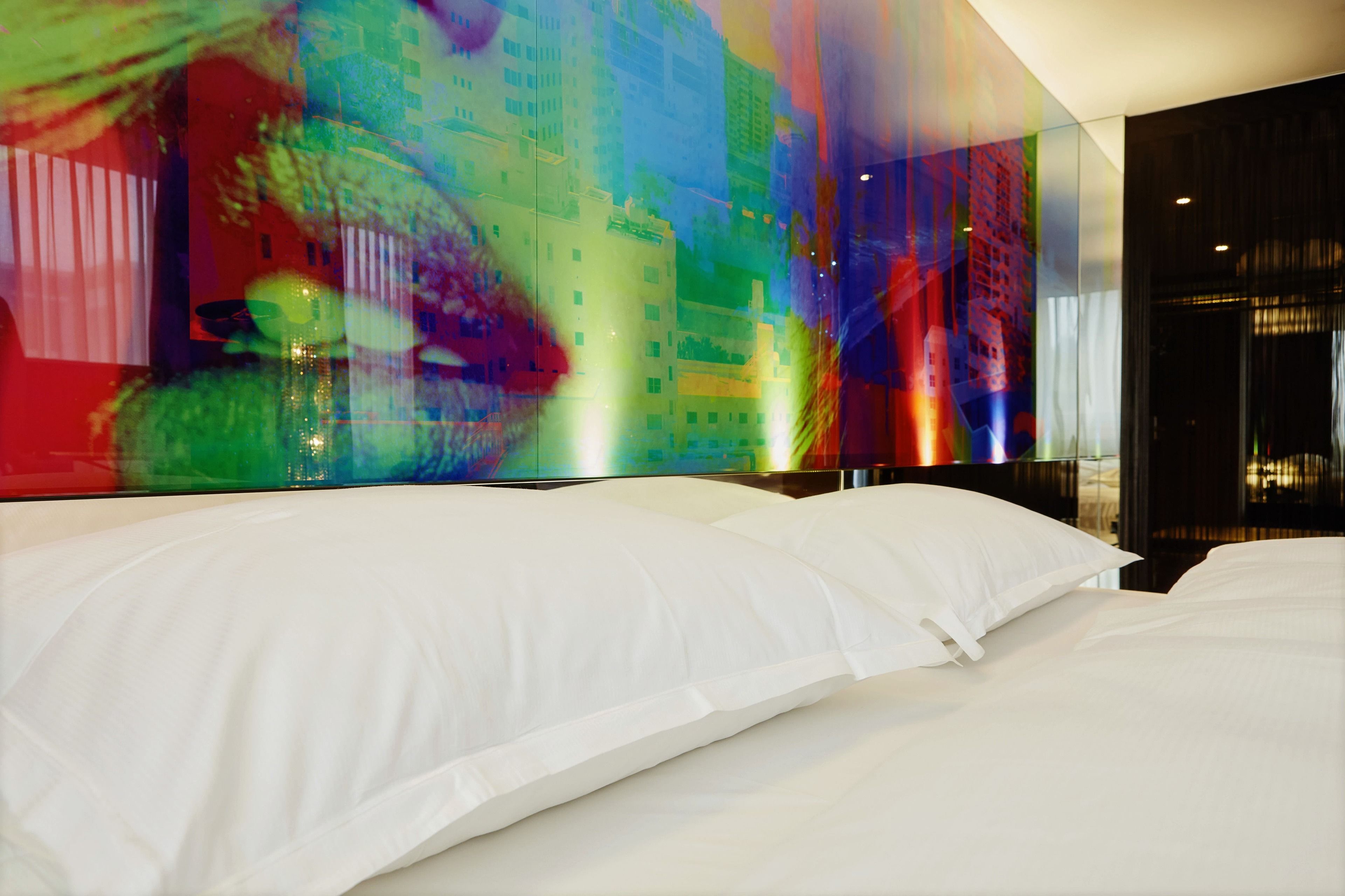 A modern hotel room with a large bed, a glass wall, and a colorful abstract artwork.