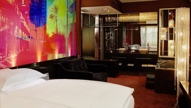 Een moderne hotelkamer met een groot bed, een zithoek met een zwarte bank, een grote spiegel en levendige abstracte wanddecoratie.