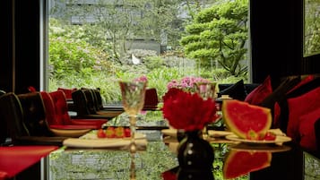 Un comedor con vistas a un jardín a través de un ventanal grande, con una mesa dispuesta con platos, un florero con flores y un vaso de agua.