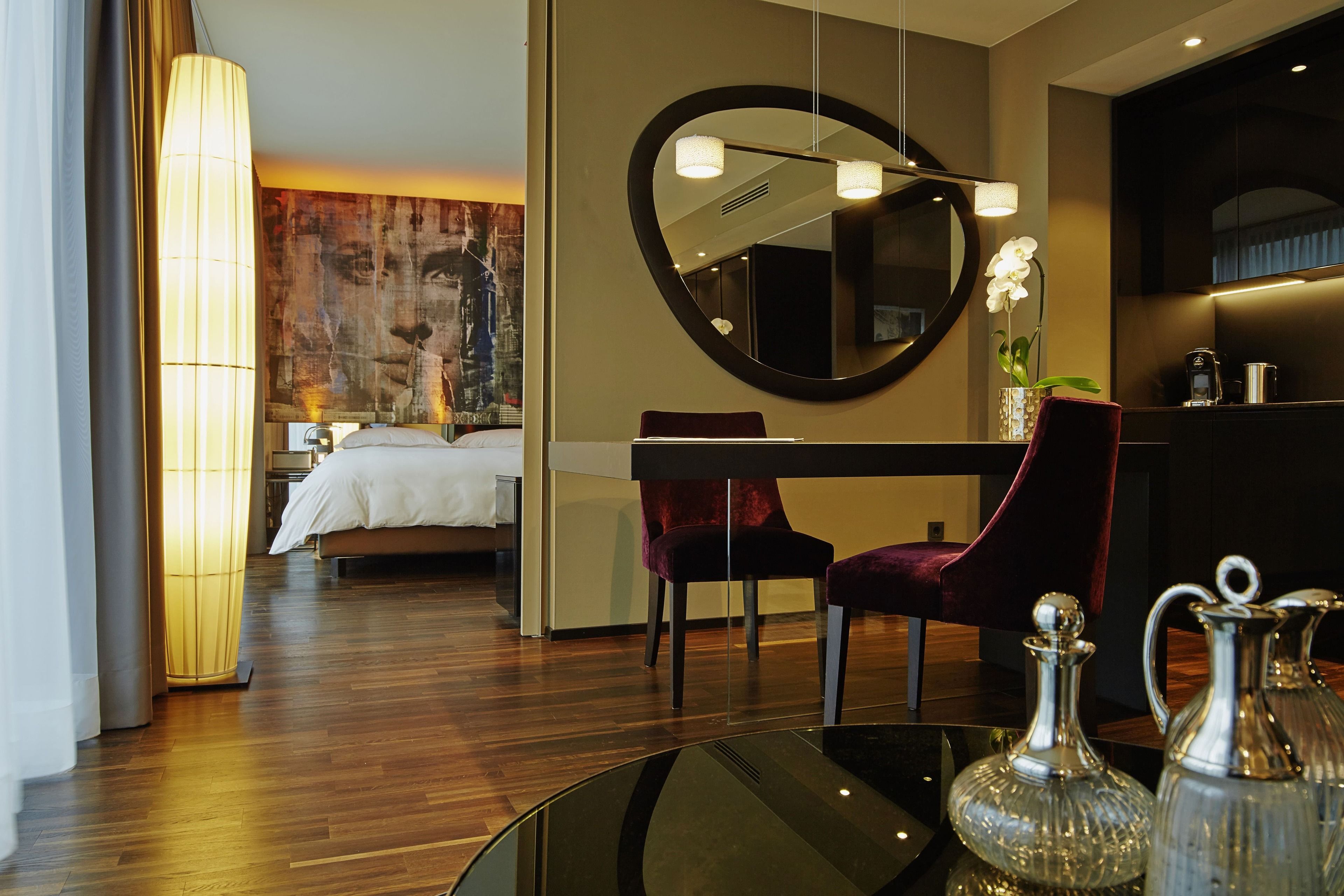 A modern hotel room with a large bed, a sitting area with a chair, a round mirror, and a table with decorative items.