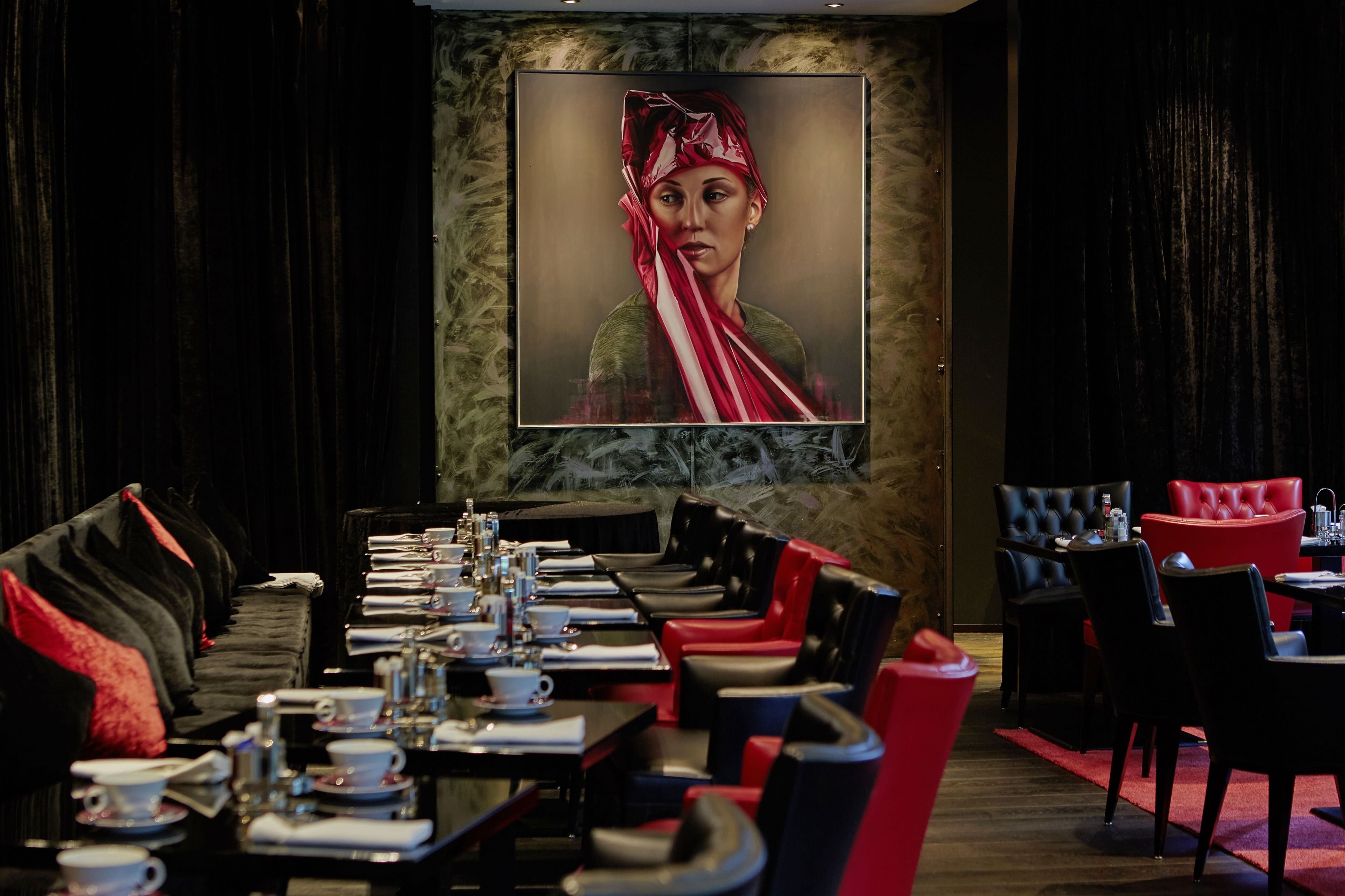 A restaurant with a long table set for dining, black and red upholstered chairs, and a large painting on the wall.