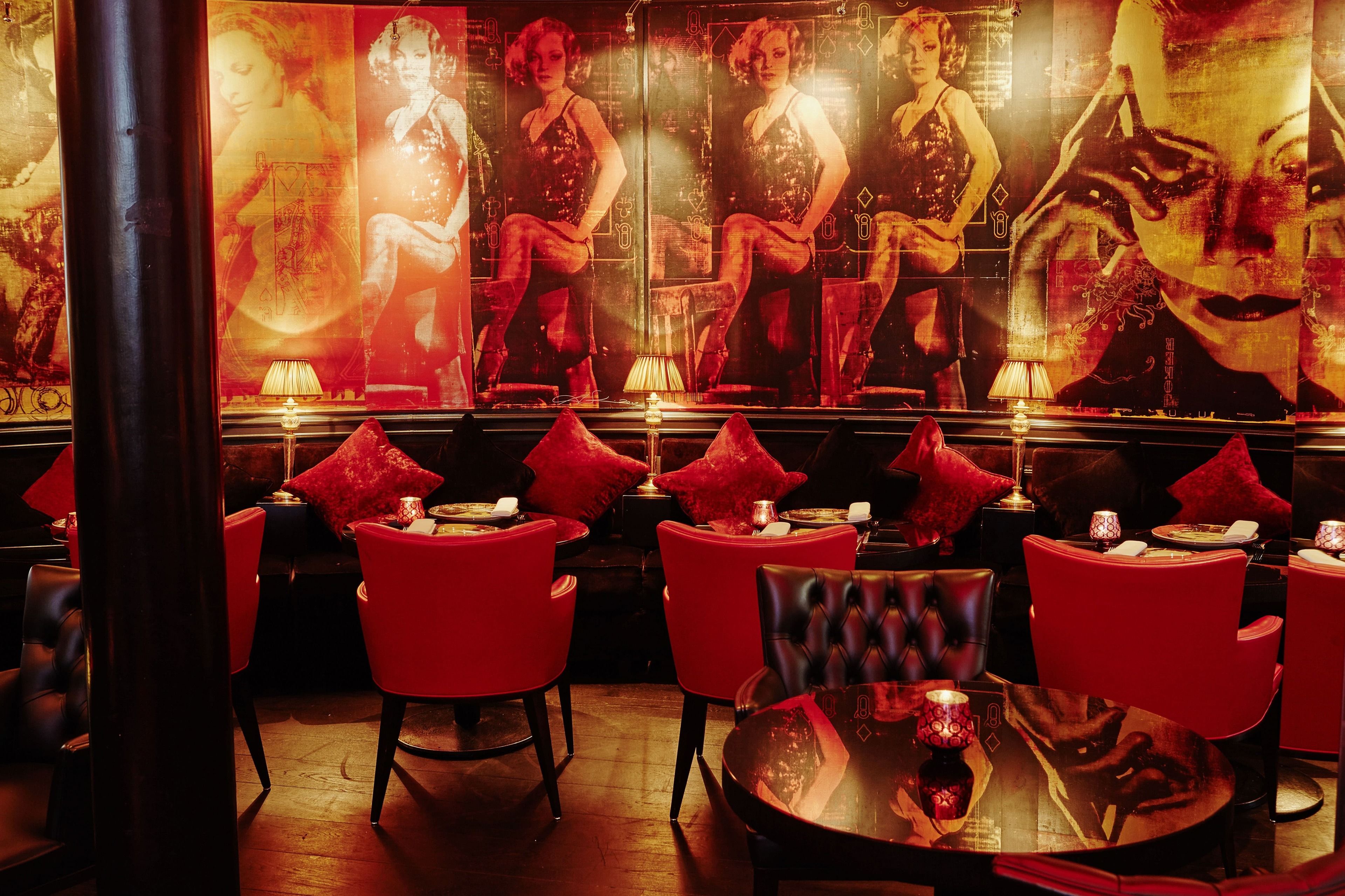 A dimly lit bar area with red upholstered chairs and tables, featuring posters of women in evening attire.