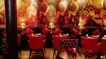 A dimly lit bar area with red upholstered chairs and tables, featuring posters of women in evening attire.