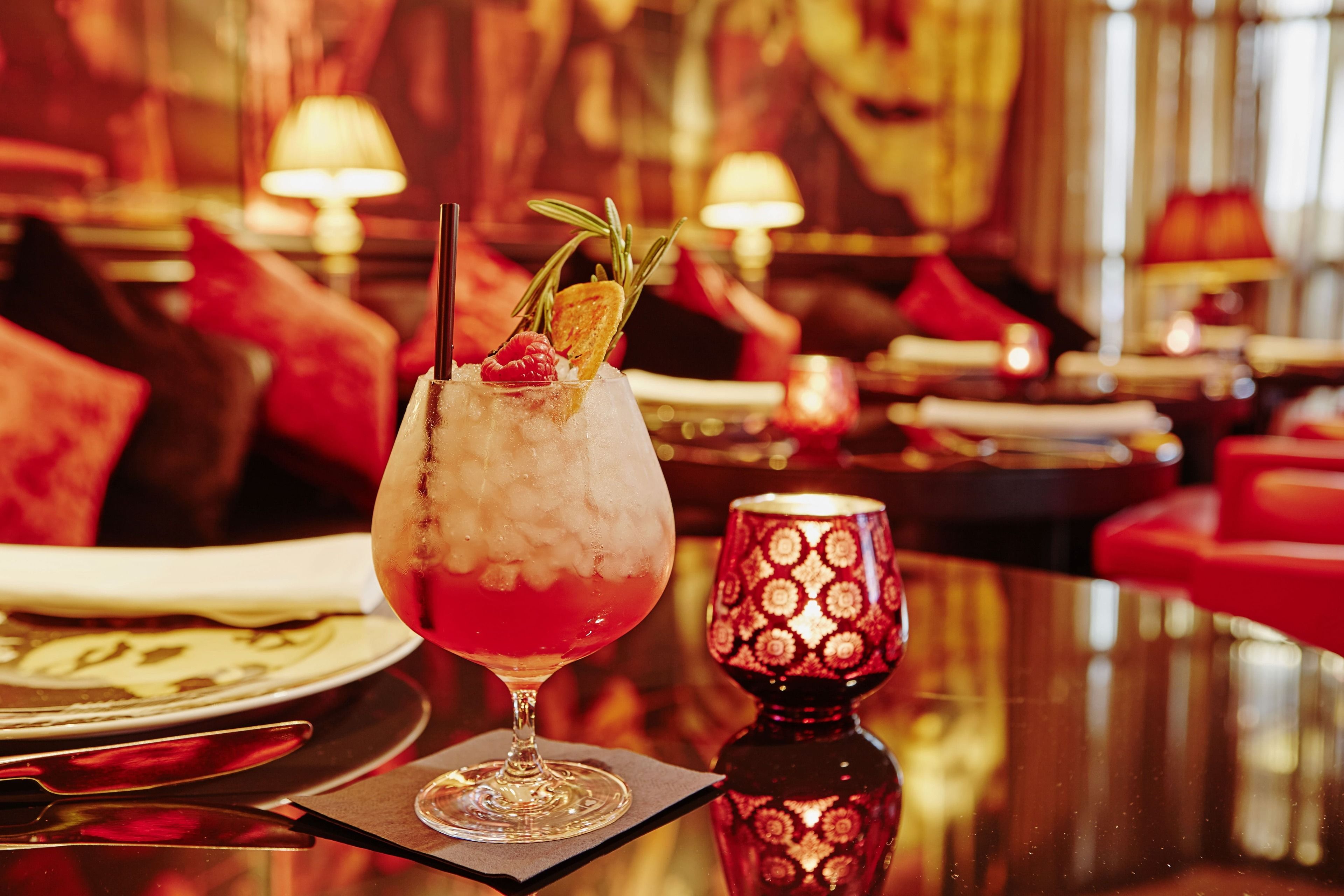 Ein Cocktail mit Eis und Garnitur in einem Glas, eine Kerze und ein Teller mit Serviette auf einem Tisch in einem gemütlich beleuchteten Raum.