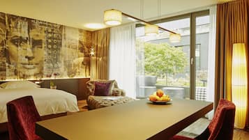 A hotel room with a large bed, a dining table with chairs, a balcony with a view of greenery, and a wall with a collage of images.