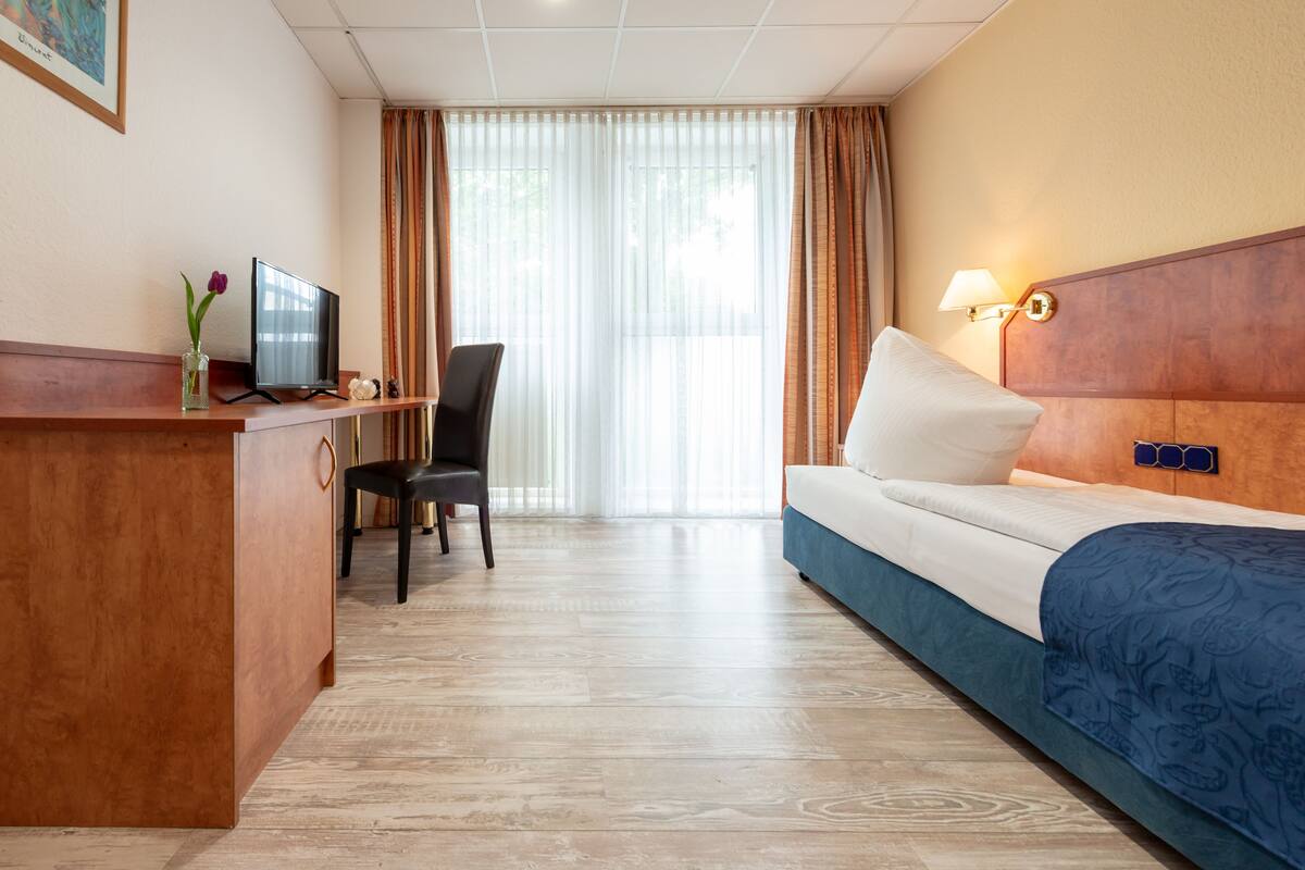 A hotel room with a wooden desk, a television, a chair, a bed with a blue bedspread, and a window with curtains.